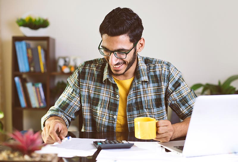 A man in a flannel shirt drinking a cup of coffee looking over some papers next to his laptop